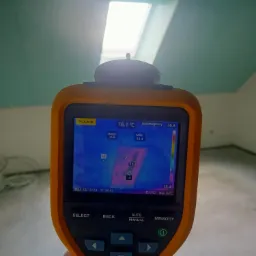 Pomiar temperatury w pomieszczeniu za pomocą kamery termowizyjnej Fluke, widoczny ekran urządzenia z kolorowym rozkładem temperatur na tle surowego, jasnego pomieszczenia z oknem dachowym.