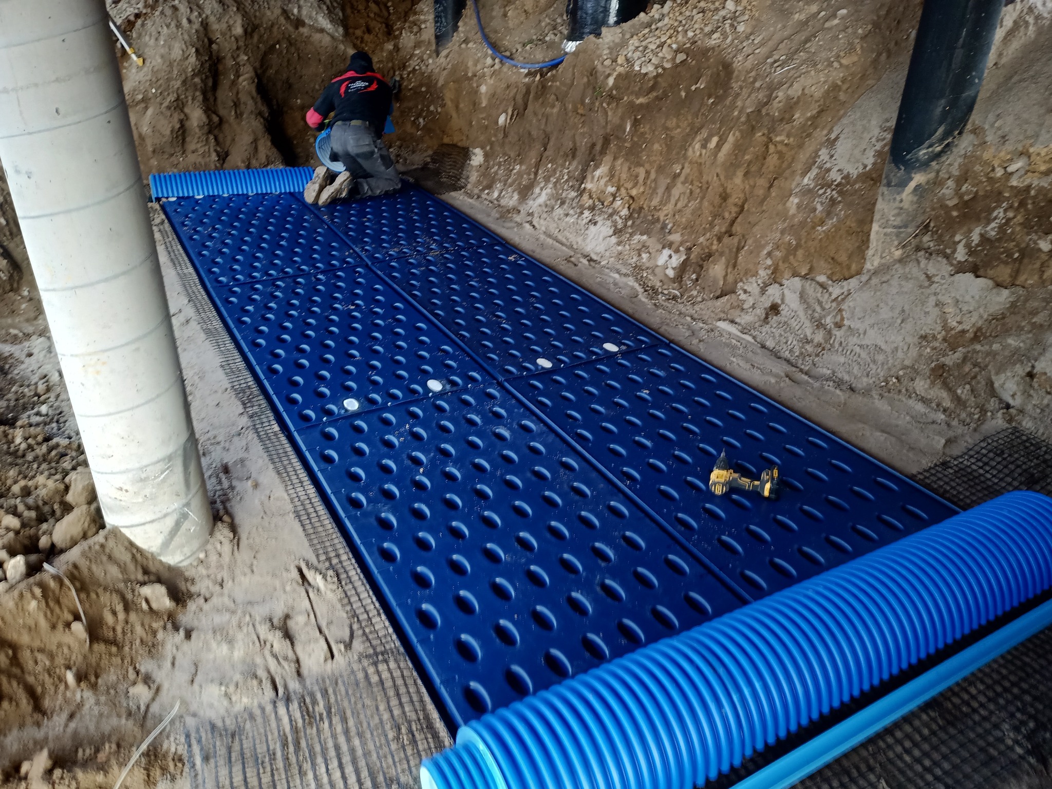 Instalacja drenażu: niebieskie panele drenażowe ułożone na warstwie geowłókniny w wykopie, pracownik w tle, widoczna wiertarka akumulatorowa.