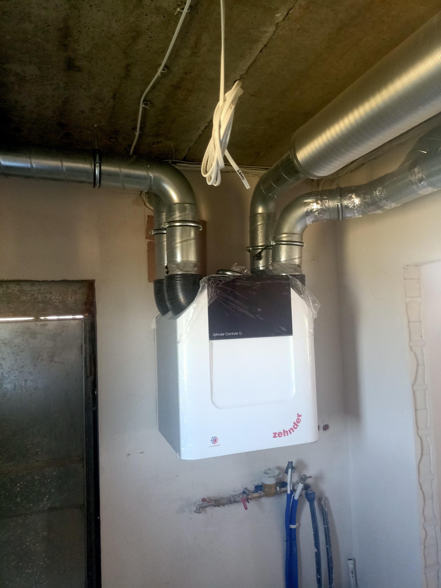 Centrala rekuperacyjna Zehnder ComfoAir Q z podłączonymi przewodami wentylacyjnymi w surowym pomieszczeniu, z widocznymi rurami instalacji wodnej i fragmentem metalowych drzwi.