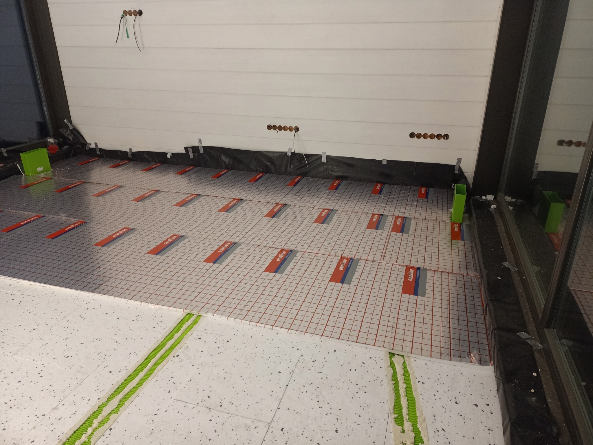 Przygotowanie podłoża pod ogrzewanie podłogowe, widoczne płyty izolacyjne z siatką i oznaczeniami producenta, zielona taśma dylatacyjna przy ścianie, przygotowane otwory w ścianie.