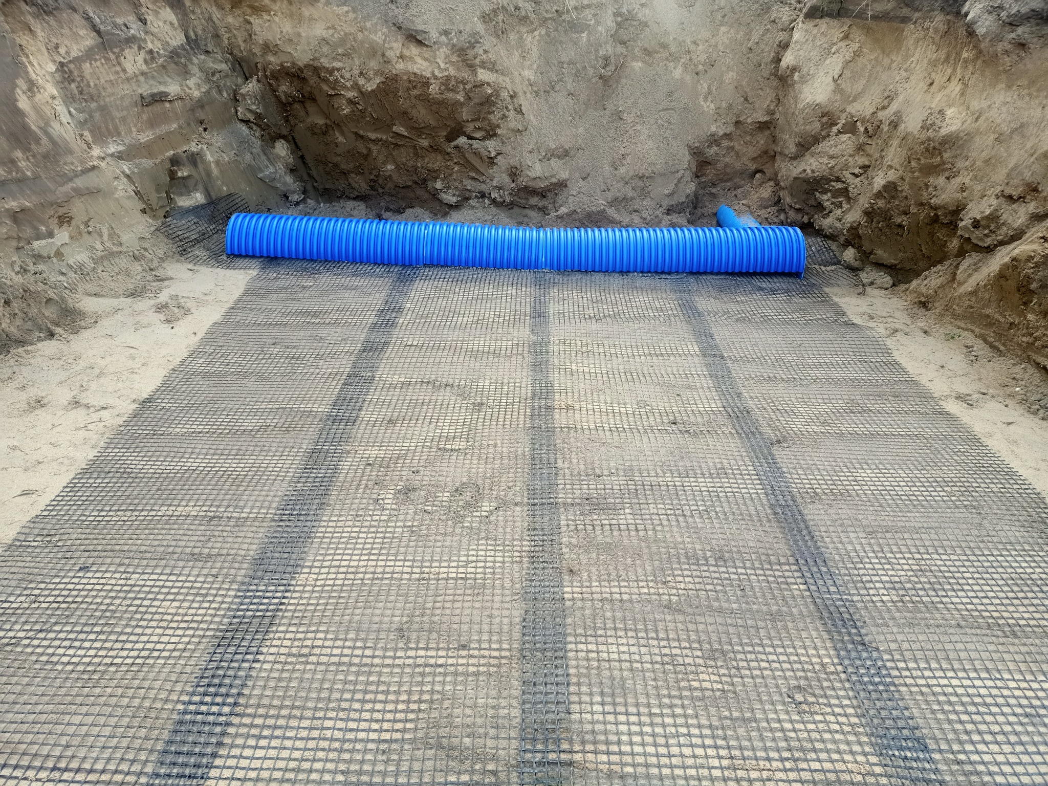 Wykonany wykop z ułożoną geokratą i niebieską rurą drenarską karbowaną na dnie, widoczne ślady po pracy koparki.
