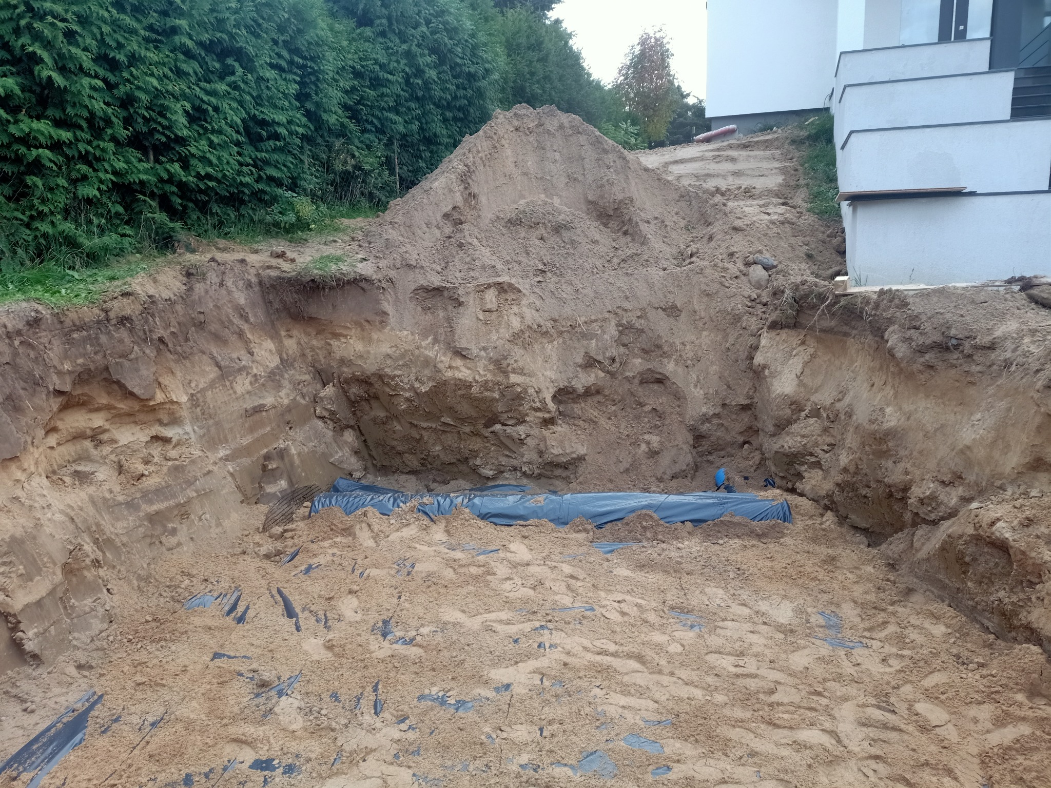 Wykop pod budynek z widoczną izolacją w postaci niebieskiej folii, pryzma ziemi obok budynku z białymi schodami.