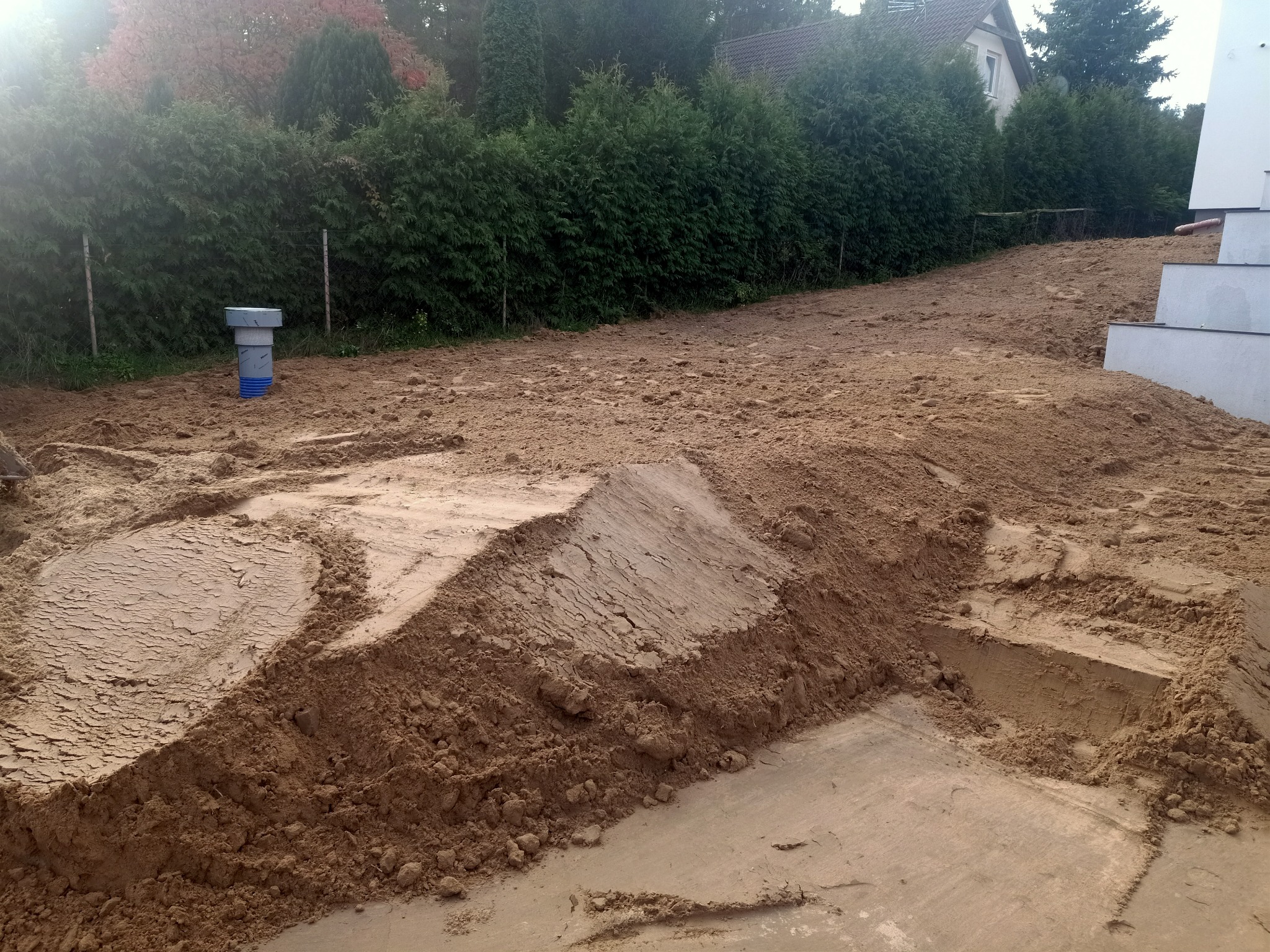 Przygotowanie terenu pod budowę domu jednorodzinnego: wyrównany teren z hałdami ziemi, widoczna studzienka kanalizacyjna i schody wejściowe do budynku.