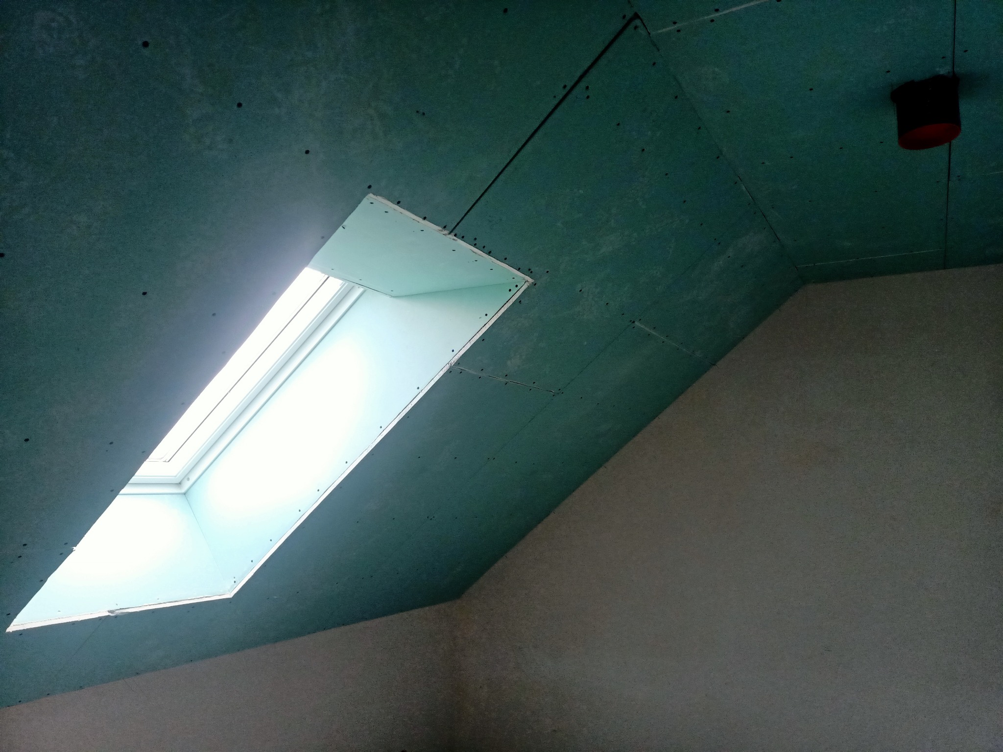 Montaż okna dachowego w skośnym suficie z płyt gipsowo-kartonowych, widoczne łączenia płyt i otwór wentylacyjny.
