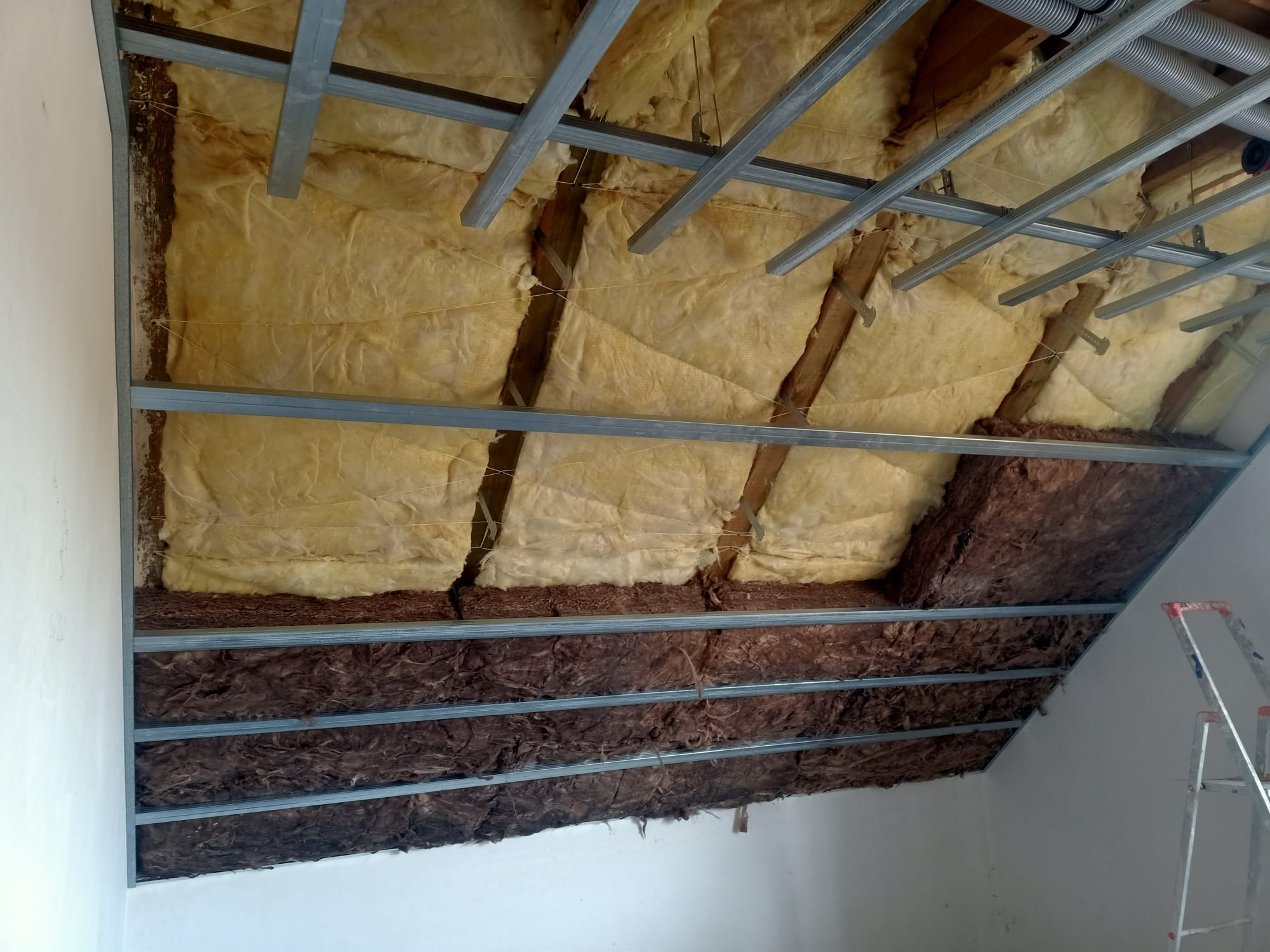 Ocieplenie poddasza wełną mineralną w dwóch warstwach, widoczne metalowe profile konstrukcyjne, drabina oparta o ścianę.