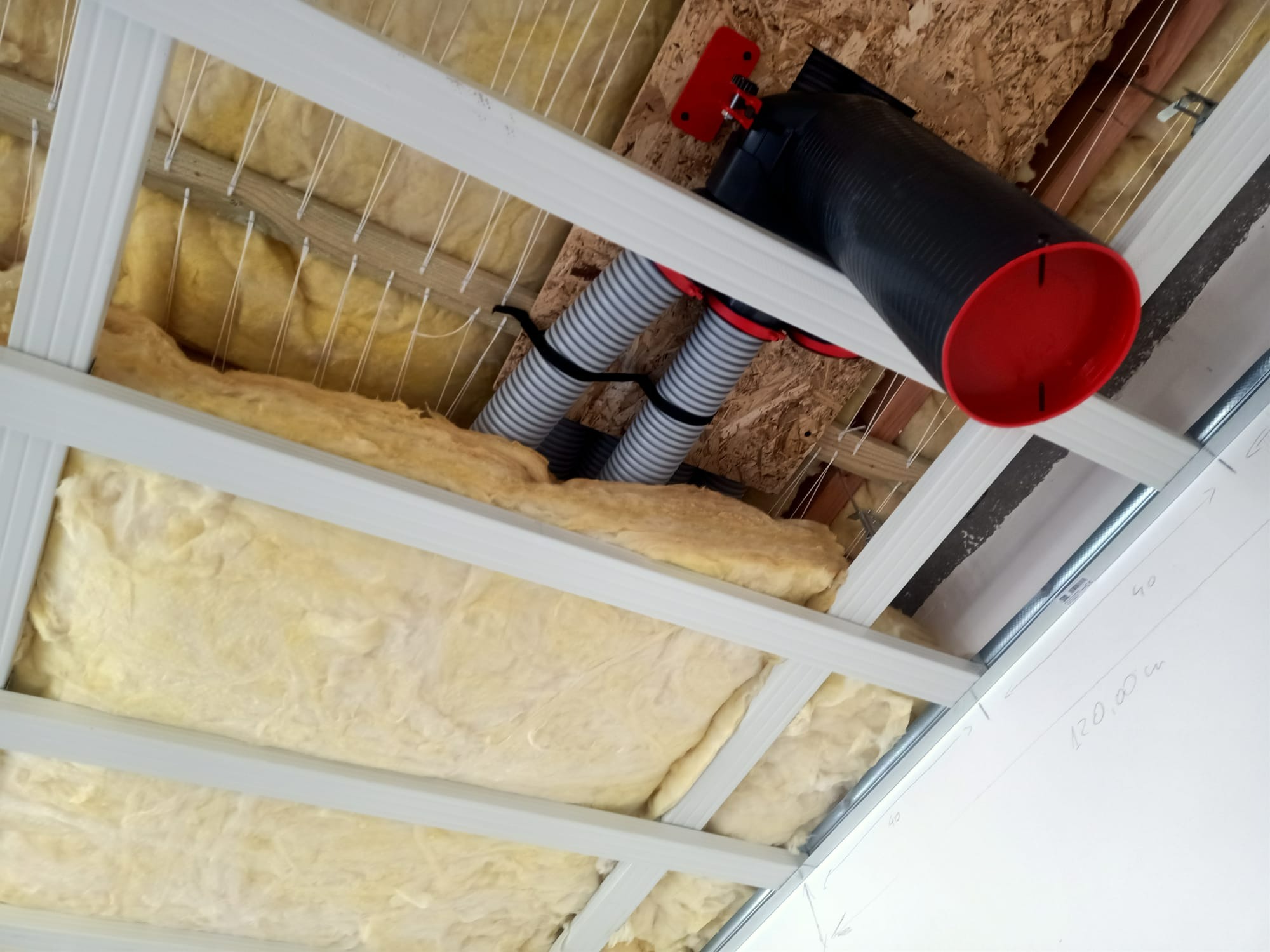 Czarna rura wentylacyjna z czerwonymi zakończeniami podwieszona do sufitu z ociepleniem z wełny mineralnej, widoczne elastyczne przewody wentylacyjne i konstrukcja nośna sufitu.