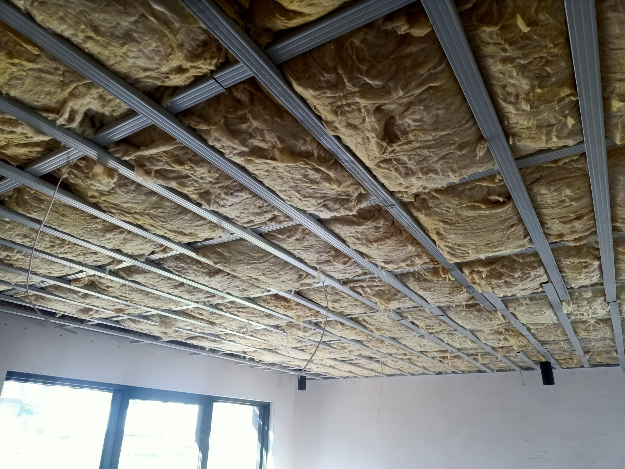 Montaż izolacji z wełny mineralnej w suficie podwieszanym z metalową konstrukcją nośną, widoczne okno i fragment ściany.