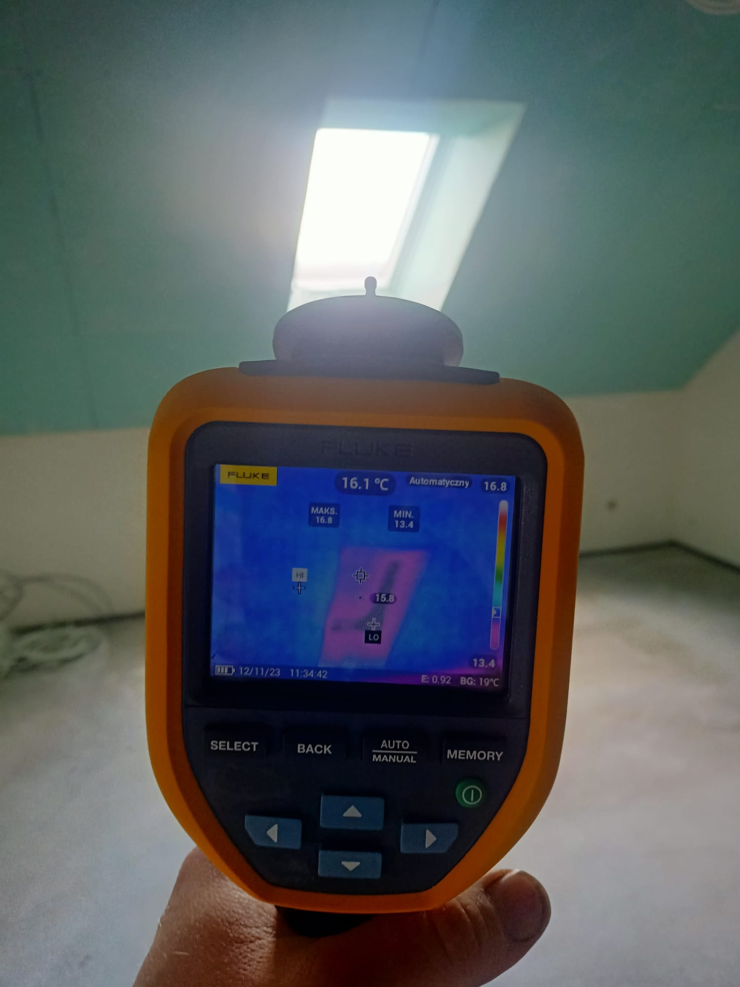 Pomiar temperatury w pomieszczeniu za pomocą kamery termowizyjnej Fluke, widoczny ekran urządzenia z kolorowym rozkładem temperatur na tle surowego, jasnego pomieszczenia z oknem dachowym.