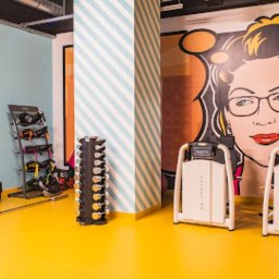 TOPAGO GROUP SPÓŁKA Z OGRANICZONĄ ODPOWIEDZIALNOŚCIĄ - Kolorowe wnętrze siłowni z grafiką kobiety w okularach, sprzętem do ćwiczeń i ścianą w paski. Żółta podłoga i różnorodne barwy ścian tworzą energiczną atmosferę.