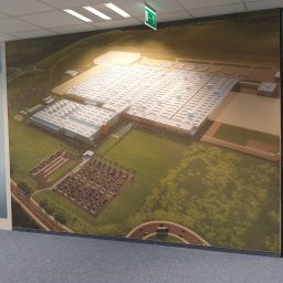 TOPAGO GROUP SPÓŁKA Z OGRANICZONĄ ODPOWIEDZIALNOŚCIĄ - Fototapeta w biurze przedstawia wizualizację fabryki z parkingiem, widok z lotu ptaka. W tle widoczne drzwi i odbicie osoby robiącej zdjęcie.