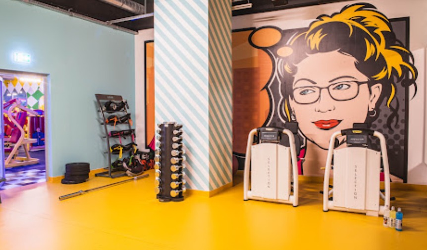 Kolorowe wnętrze siłowni z grafiką kobiety w okularach, sprzętem do ćwiczeń i ścianą w paski. Żółta podłoga i różnorodne barwy ścian tworzą energiczną atmosferę.