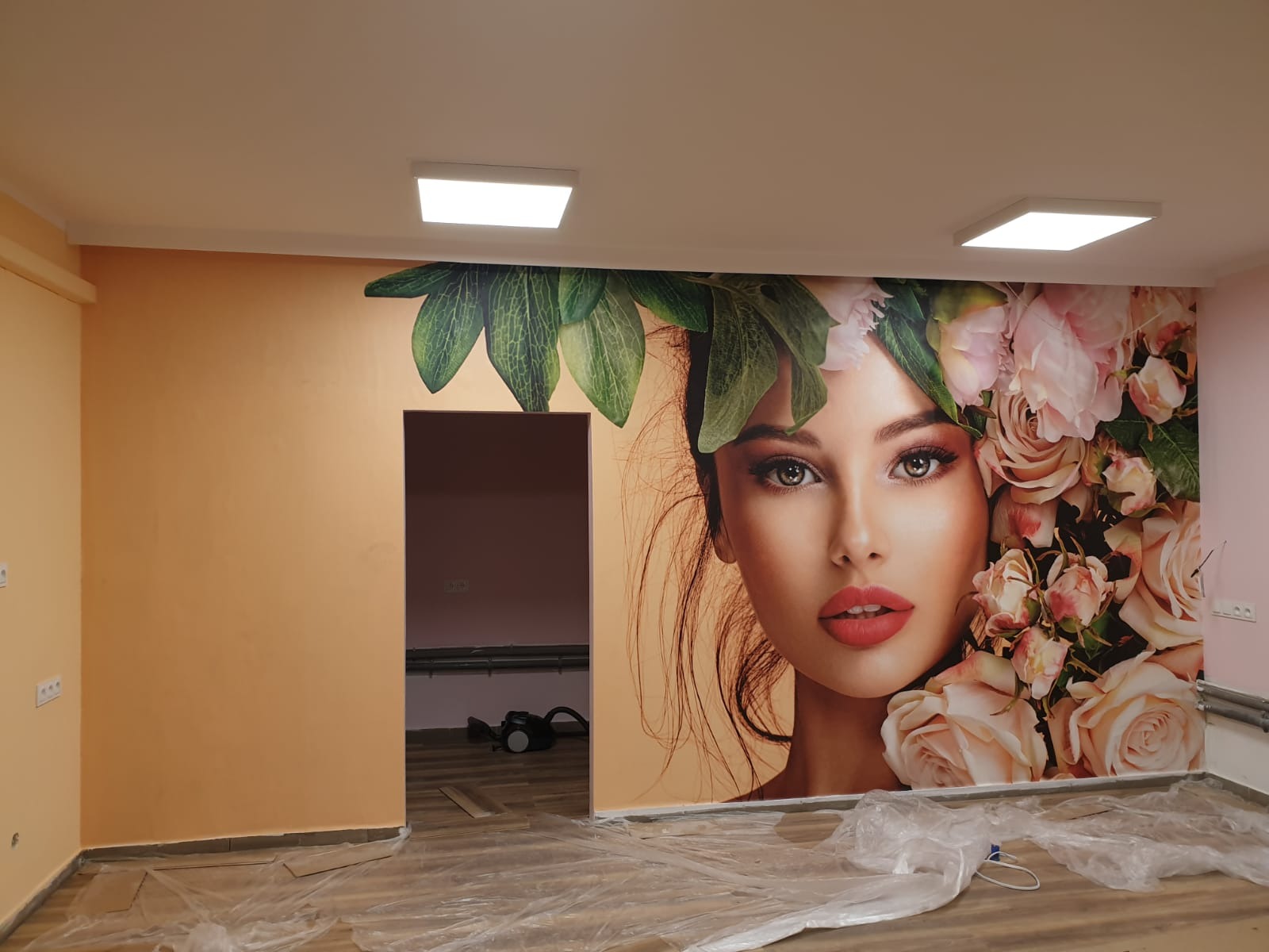 Fototapeta z wizerunkiem kobiety w kwiatach na ścianie w pokoju, zabezpieczona folią podłoga, widoczne oświetlenie sufitowe.