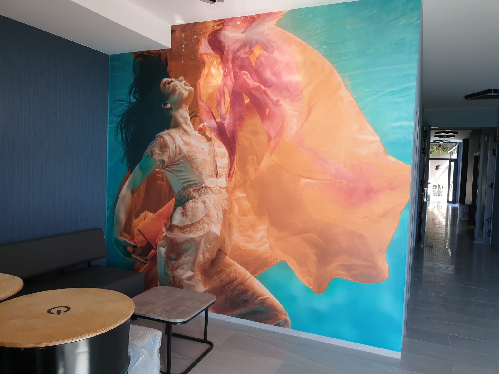 Fototapeta z motywem podwodnym przedstawiająca kobietę w zwiewnej sukni, umieszczona na ścianie w nowoczesnym wnętrzu z szarą sofą i stolikami.