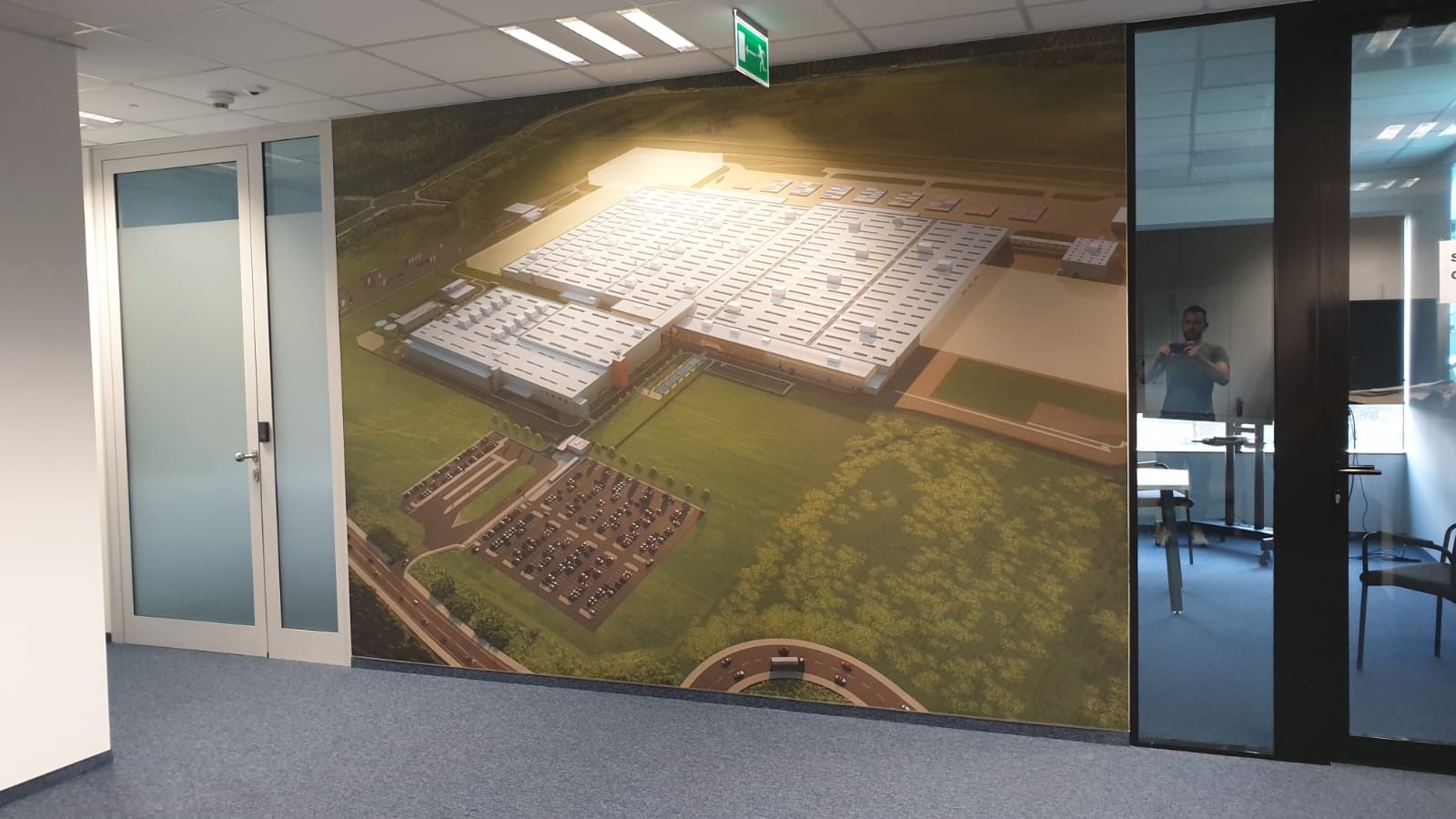 Fototapeta w biurze przedstawia wizualizację fabryki z parkingiem, widok z lotu ptaka. W tle widoczne drzwi i odbicie osoby robiącej zdjęcie.