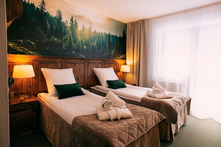 Pokój hotelowy z dwoma łóżkami, drewnianym zagłówkiem i fototapetą z motywem lasu. Przy oknie zwiewne zasłony, a na łóżkach ręczniki i szlafroki.