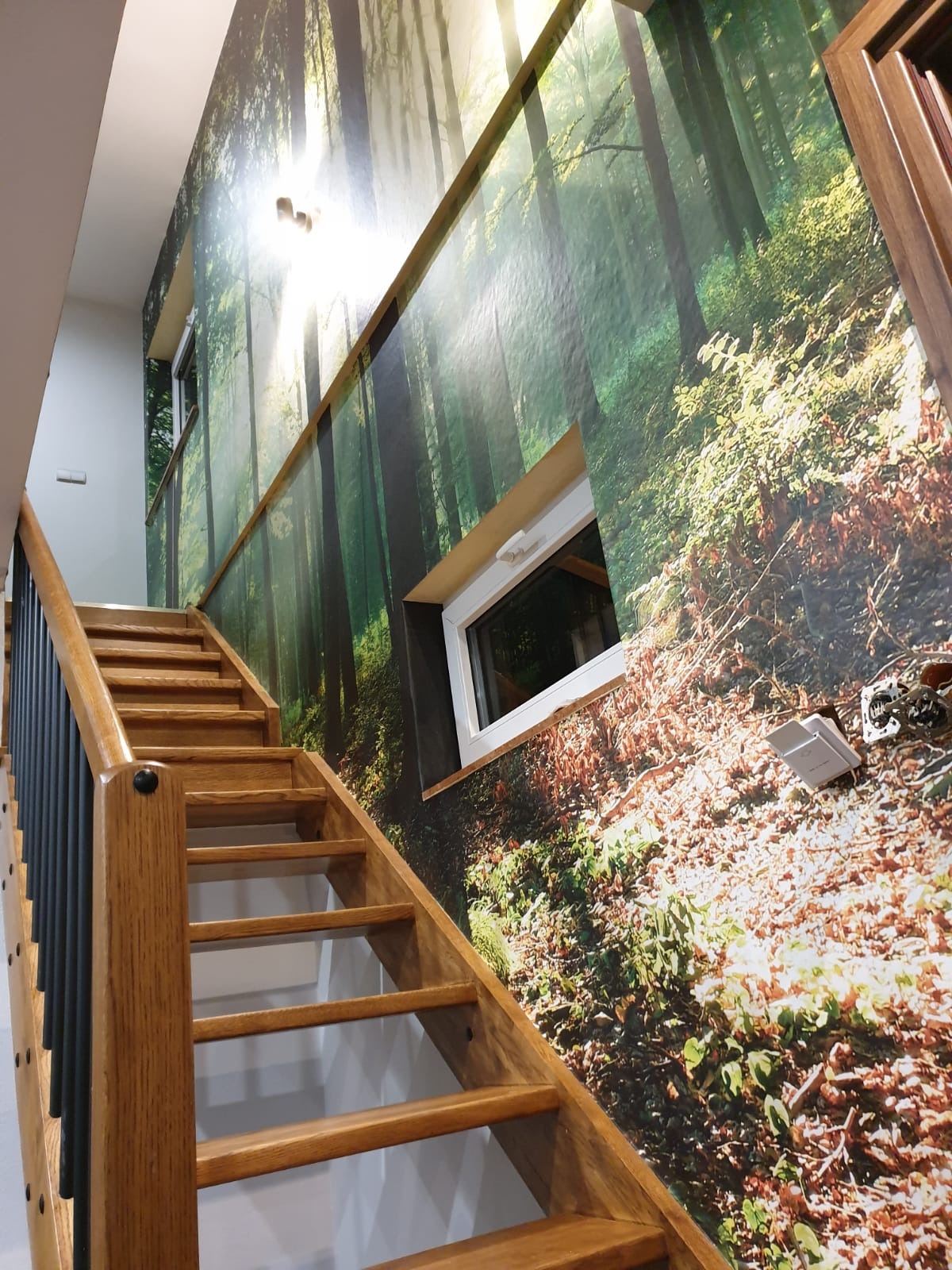 Ściana w klatce schodowej z fototapetą leśną, drewniane schody z czarną balustradą. Okno wkomponowane w leśny motyw. Widoczne gniazdko elektryczne.