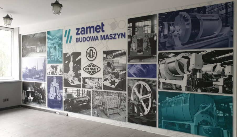 Ściana z fototapetą prezentującą kolaż zdjęć maszyn i logo firmy ZAMET, z przewagą kolorów szarości i błękitu, w jasnym pomieszczeniu z oknem.