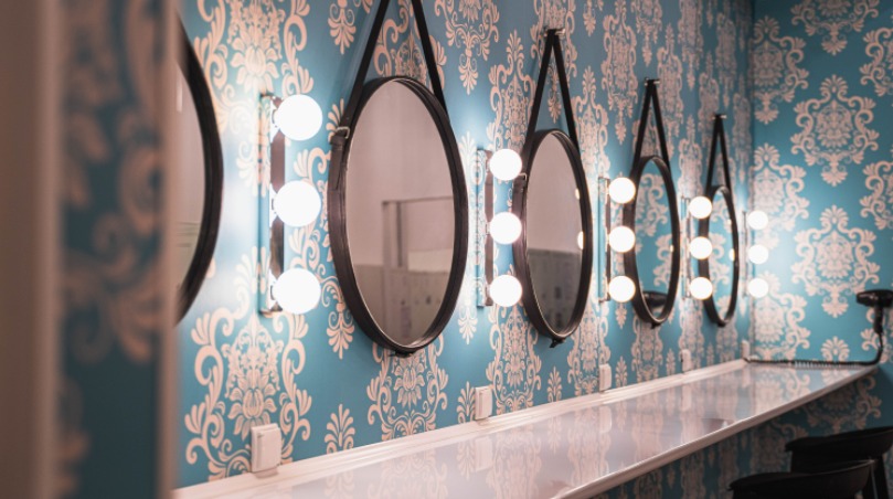 Niebieska ściana w białe wzory, z rzędem okrągłych luster z oświetleniem, wiszących na czarnych paskach. Długi, biały blat pod lustrami.