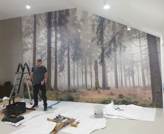 Montaż fototapety z motywem leśnym na ścianie skośnej. Widoczny pracownik, drabina i narzędzia na podłodze zabezpieczonej folią malarską.