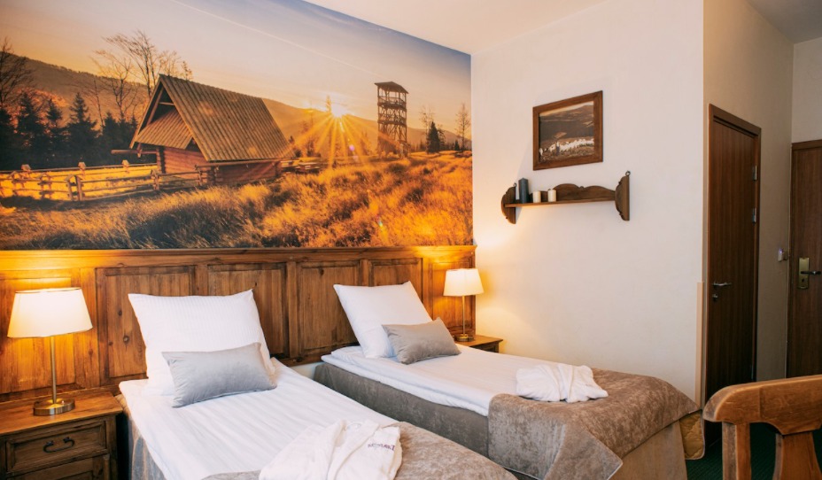 Pokój hotelowy z dwoma łóżkami pojedynczymi i fototapetą górskiego krajobrazu nad wezgłowiem. Drewniane meble, lampki nocne i dekoracje.