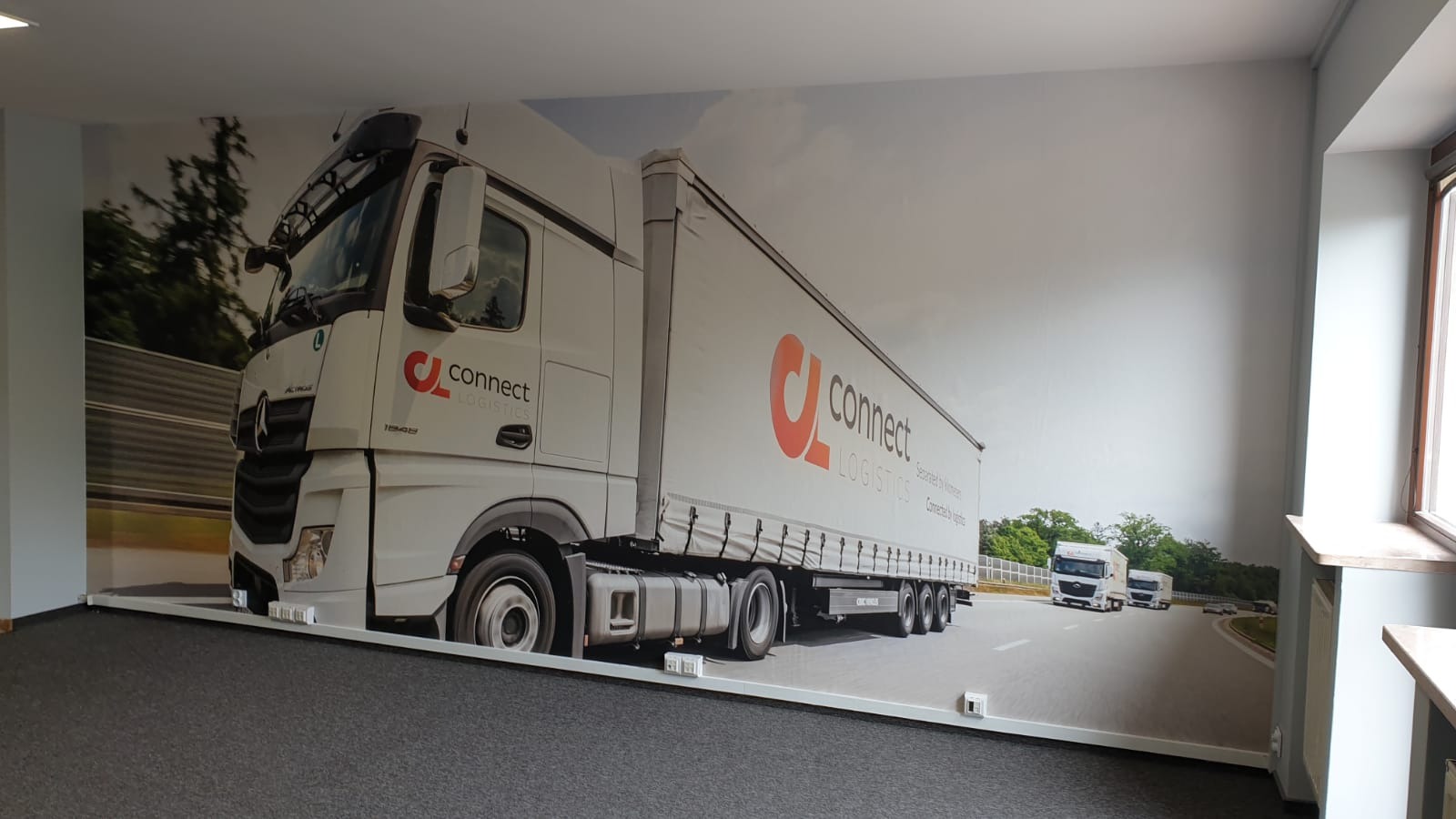 Wnętrze biura z fototapetą na ścianie, przedstawiającą białą ciężarówkę firmy Connect Logistics na drodze. Widoczne gniazdka elektryczne na dole ściany.