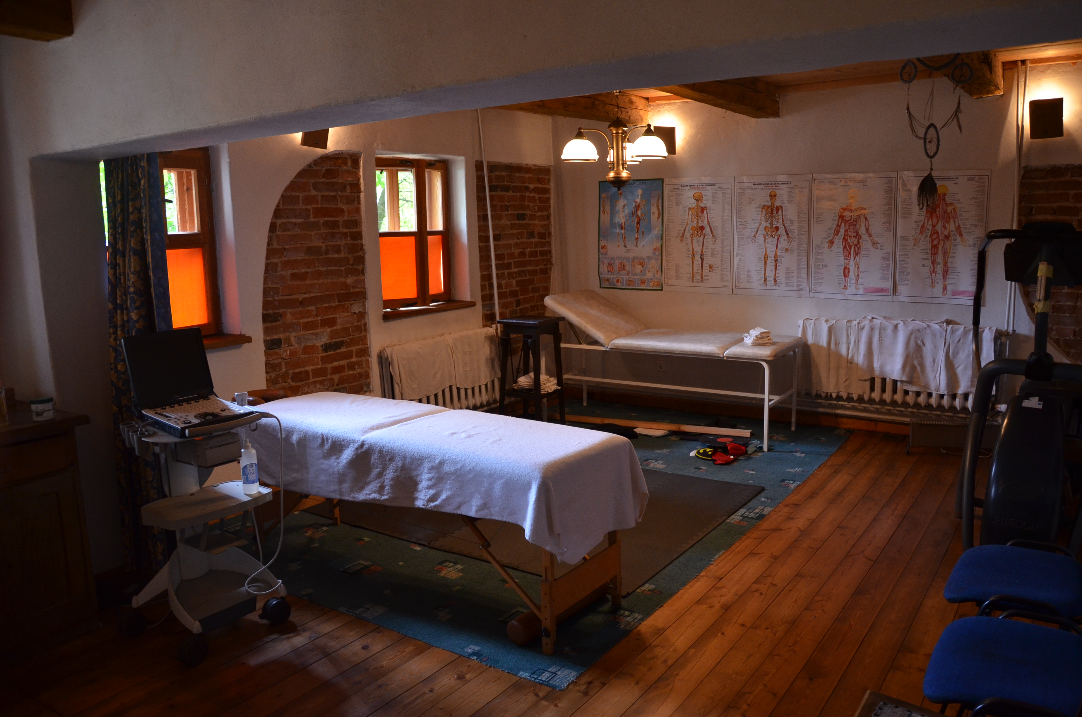 Gabinet z dwoma leżankami przykrytymi białymi ręcznikami, sprzęt do fizykoterapii, plakaty z układem kostnym, drewniana podłoga i ceglane ściany z oknami.
