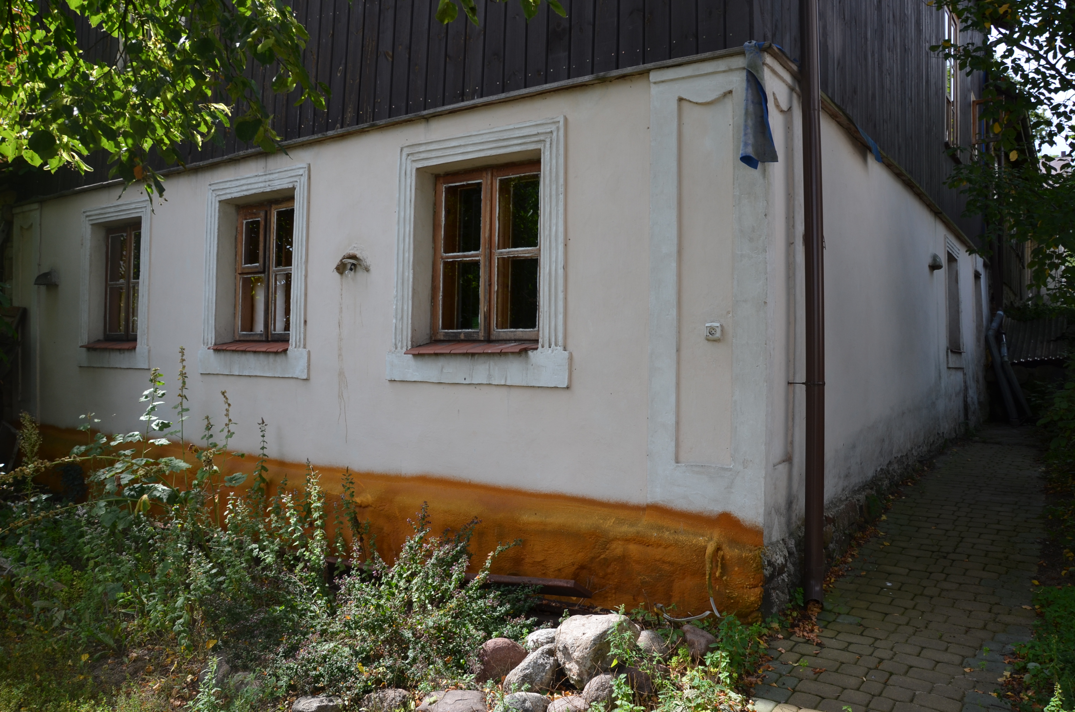 Elewacja budynku z oknami, z widocznymi śladami wilgoci i roślinnością u podstawy, z narożną rynną i fragmentem chodnika z kostki.
