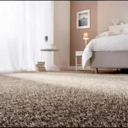 Beżowy dywan shaggy w sypialni z łóżkiem, stolikiem nocnym, lampką i zasłonami.