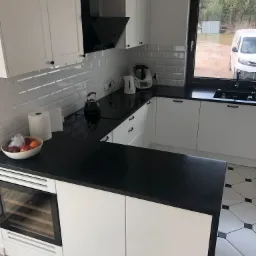 Nowoczesna, biała kuchnia z czarnym blatem i płytkami typu metro, wyposażona w ekspres do kawy, czajnik i wbudowaną chłodziarkę do wina. Na blacie misa z owocami i rolka ręcznika papierowego. Widok...