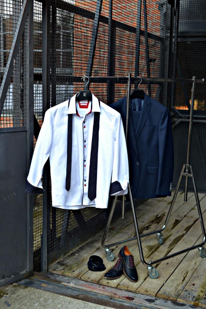 Stylizacja męska: biała koszula z czarnymi paskami i czerwonymi akcentami, granatowy garnitur, skórzane buty z czerwonymi sznurówkami i pasek na wieszaku na kółkach, na tle metalowej konstrukcji...