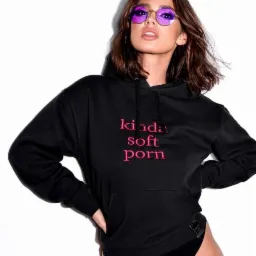 Kobieta w czarnej bluzie z kapturem i majtkach z haftowanym napisem 'kinda soft porn', nosząca futurystyczne okulary z fioletowymi szkłami, pozuje na białym tle.