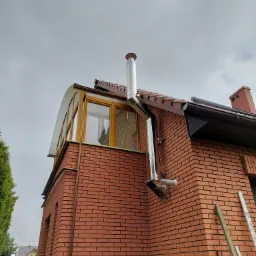 Zewnętrzny komin ze stali nierdzewnej zamontowany na ceglanym budynku z dobudowanym przeszklonym balkonem, widoczne elementy instalacji i rury odprowadzające spaliny.