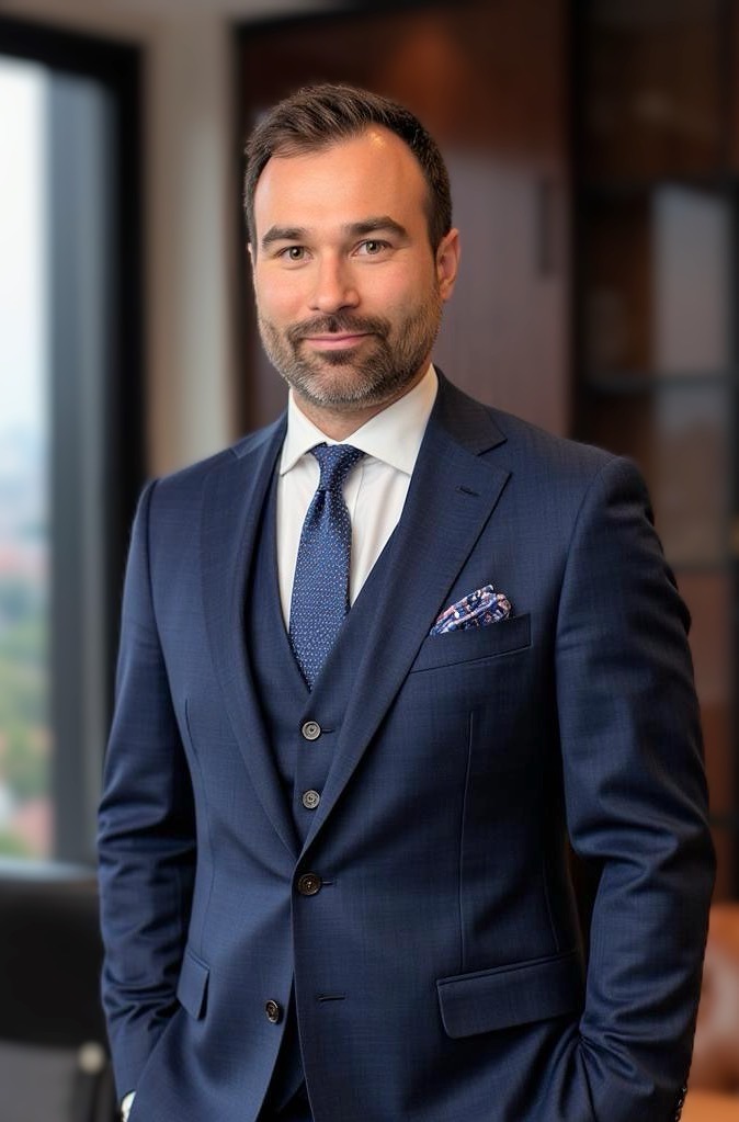 Portret adwokata w granatowym garniturze trzyczęściowym z poszetką, stojącego na tle okna i regału. Uśmiechnięty mężczyzna, profesjonalny wizerunek.