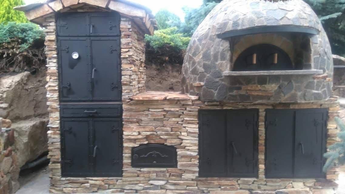 Zewnętrzny piec chlebowy z kamienia z czarnymi, metalowymi drzwiczkami, wbudowany w konstrukcję z kamienia łupanego, obok dodatkowa szafka z drzwiczkami, również z kamienia i czarnego metalu.