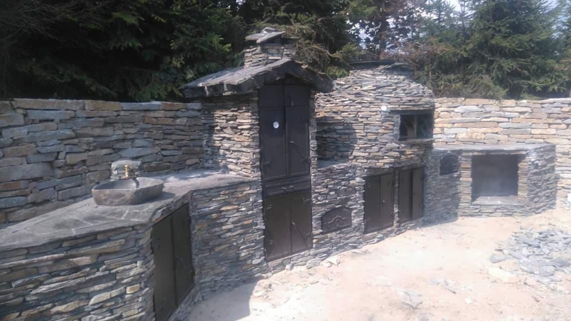 Kamienna zabudowa zewnętrzna z piecem, szafkami i zlewem, wykonana z łupka, z czarnymi, metalowymi drzwiczkami, w tle zielone drzewa.