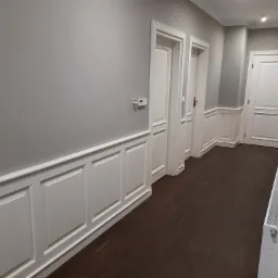 Korytarz z szarymi ścianami, ciemną drewnianą podłogą i białymi, klasycznymi drzwiami z dekoracyjnymi listwami przypodłogowymi.