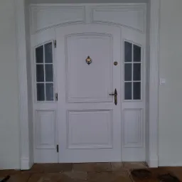 Białe drzwi wejściowe z ozdobnymi szybkami i mosiężnym wizjerem, osadzone w dopasowanej ościeżnicy z dekoracyjnymi listwami, na tle jasnej ściany i kamiennej posadzki.