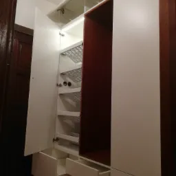 Wysoka szafa z białymi frontami, otwartymi drzwiami i szufladami, z widocznymi metalowymi półkami na buty i oświetleniem LED wewnątrz, umieszczona w pomieszczeniu z ciemnymi, drewnianymi drzwiami...