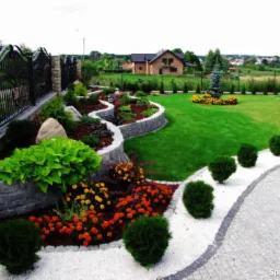 Kaskadowy ogród z kamiennymi murkami oporowymi, rabatami z kolorowymi kwiatami i krzewami, trawnikiem, ścieżką z białego żwiru i kostki brukowej, oraz widokiem na dom i okolicę w tle.