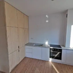 Wnętrze kuchni w trakcie wykańczania: drewniane podłogi, białe szafki z szarym blatem, zabudowa z jasnej sklejki pod sufit, niebieska ściana w tle.