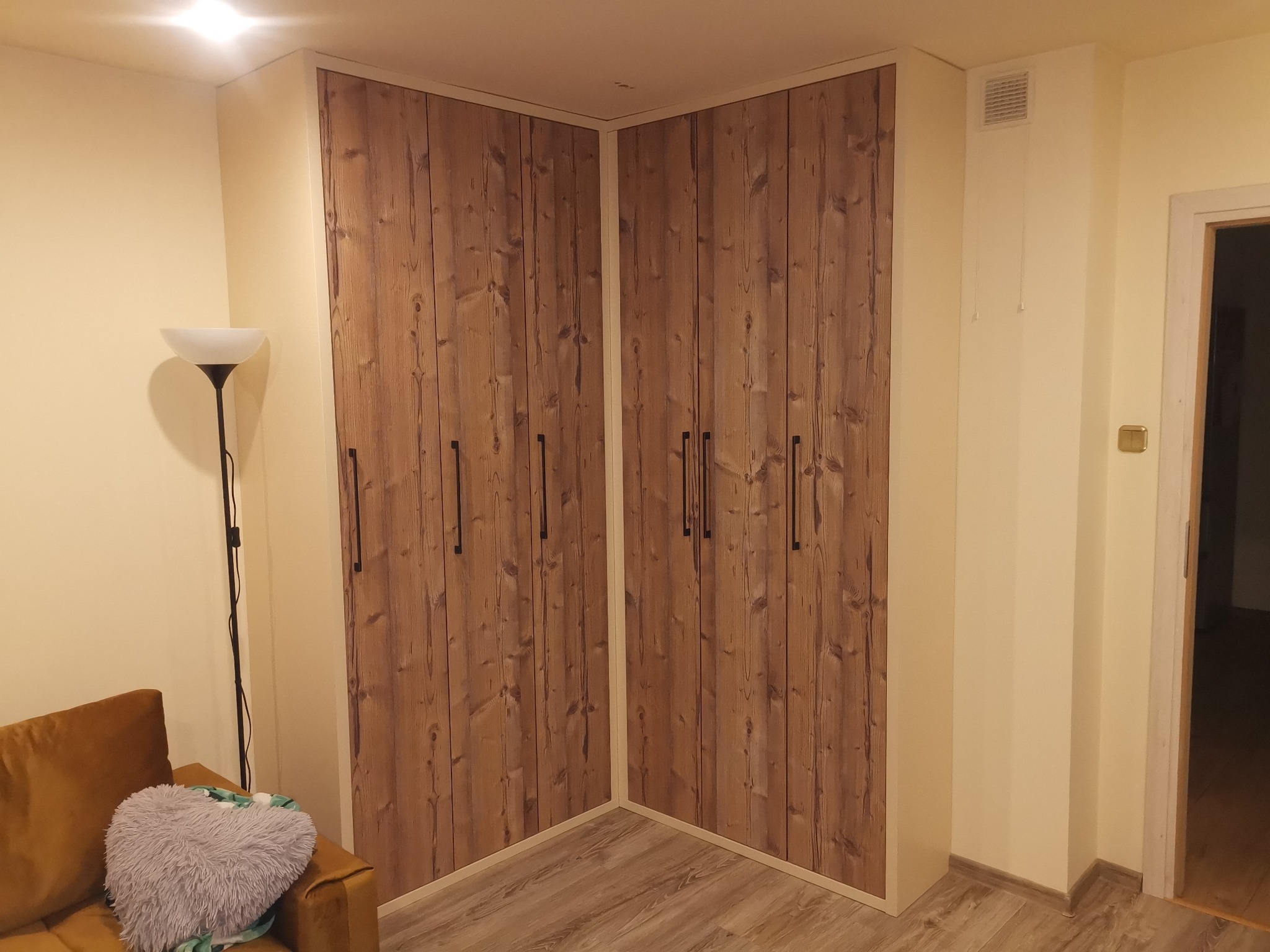 Zabudowa narożna z drewnianymi frontami szafy w pokoju, widoczny fragment sofy z poduszką w kształcie serca.