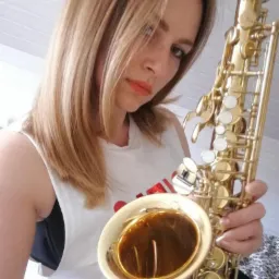 Kobieta z blond włosami trzyma saksofon, patrząc w obiektyw; widoczne odbicie światła w czarze instrumentu.