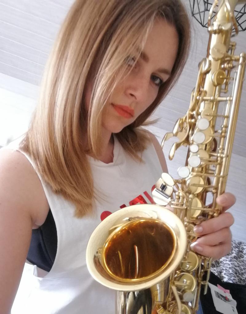 Kobieta z blond włosami trzyma saksofon, patrząc w obiektyw; widoczne odbicie światła w czarze instrumentu.