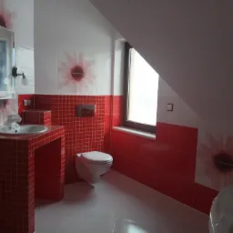 Wykończona łazienka na poddaszu z czerwono-białymi płytkami z motywem kwiatowym na ścianach, zabudowana umywalka z czerwonej mozaiki, podwieszana toaleta, okno dachowe.