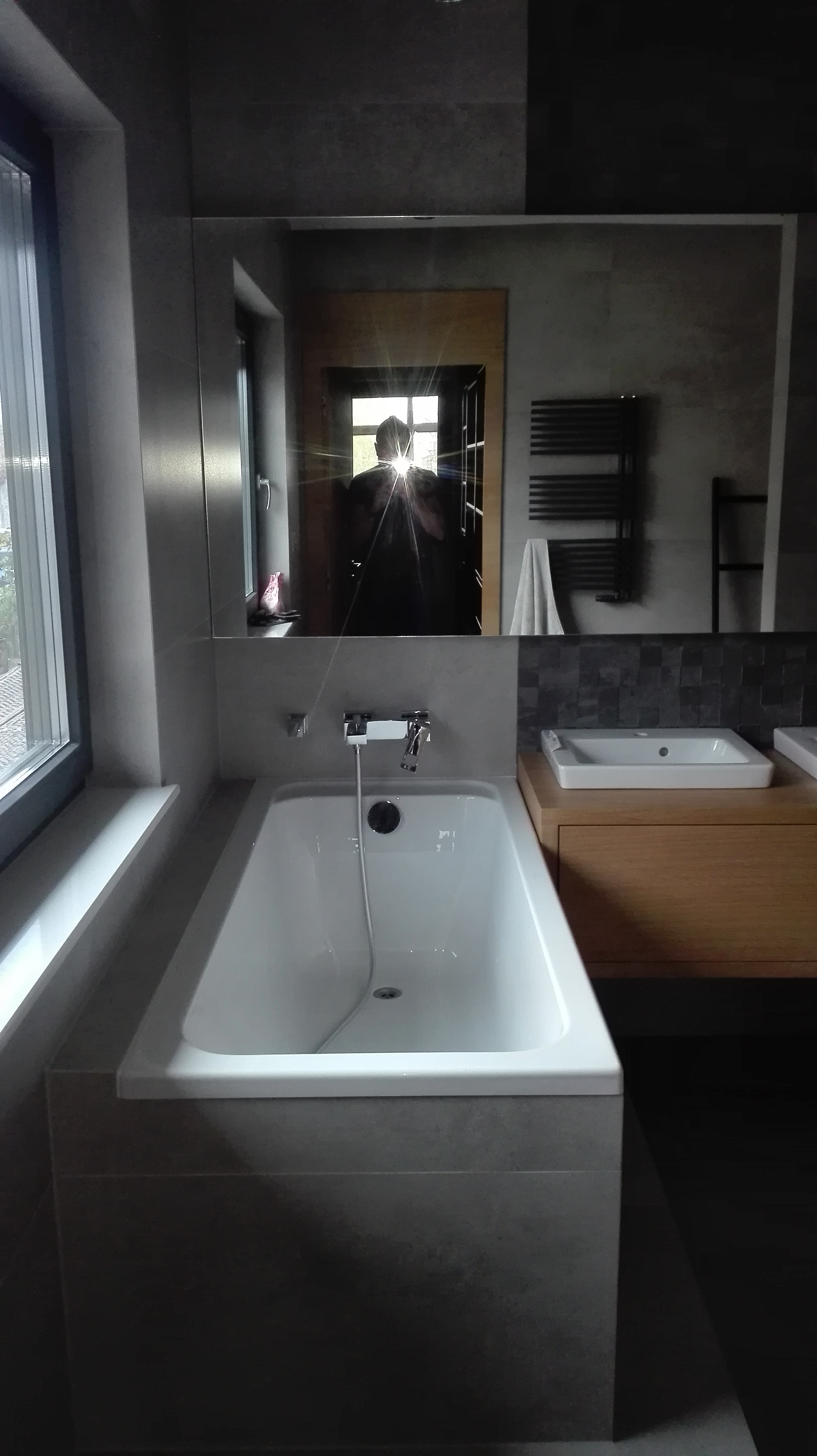 Nowoczesna łazienka z wanną obudowaną szarymi płytkami, umywalką na drewnianej szafce i dużym lustrem, w którym odbija się osoba robiąca zdjęcie.