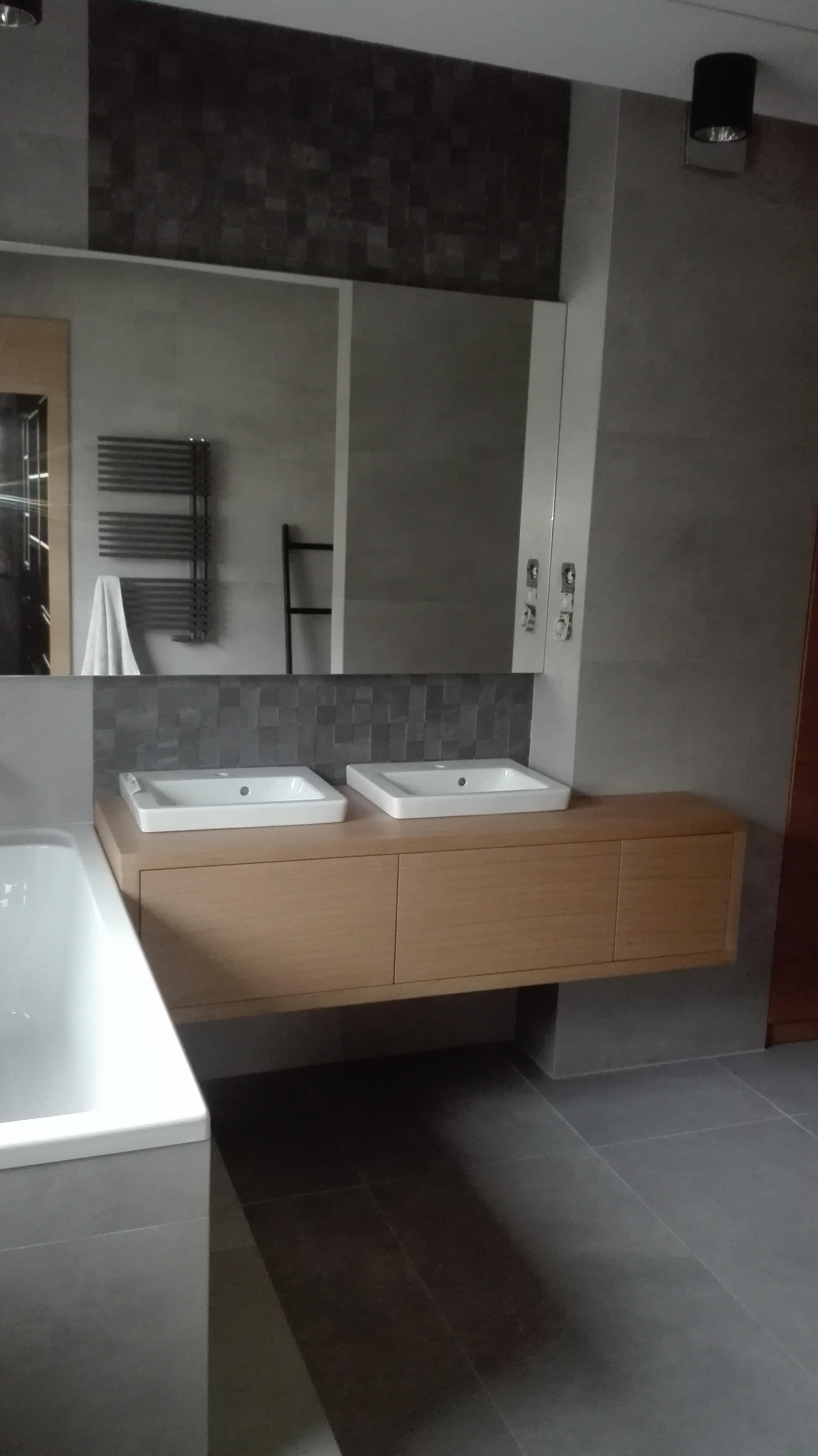 Nowoczesna łazienka z dwoma umywalkami nablatowymi na drewnianej szafce, szare płytki na ścianach i podłodze, oraz dekoracyjna mozaika nad szafką. W lustrze odbija się grzejnik drabinkowy.