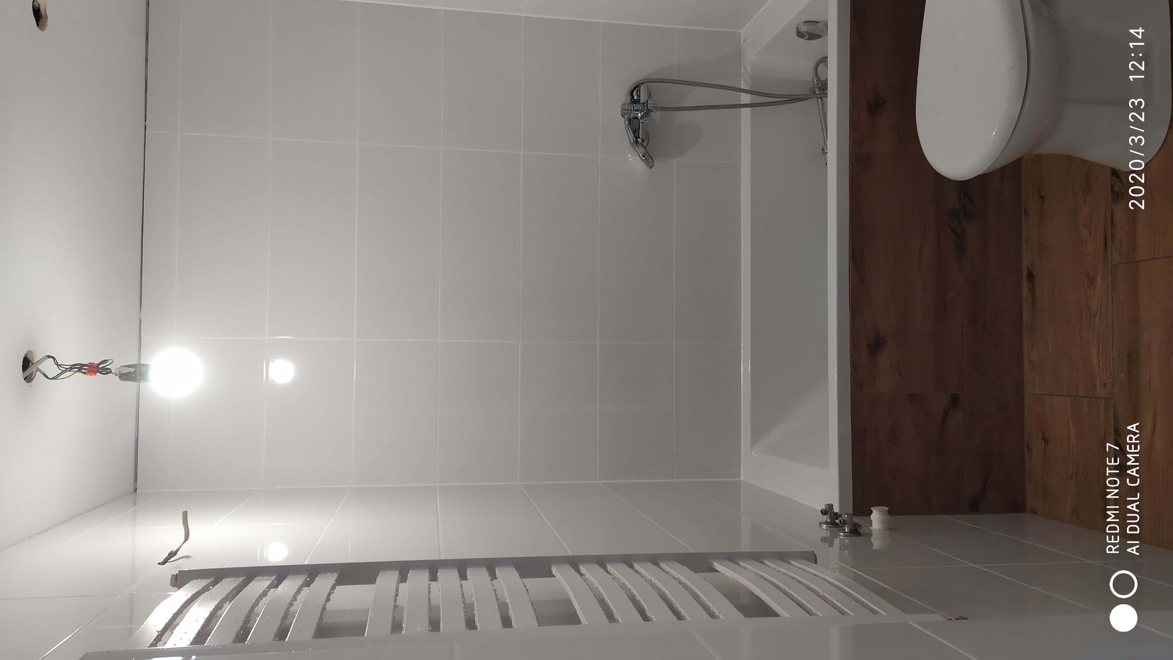 Nowa, jasna łazienka z białymi płytkami, wanną, grzejnikiem drabinkowym i fragmentem toalety, widoczne niedokończone podłączenie elektryczne.