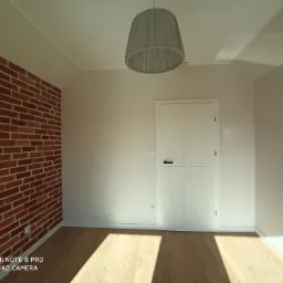 Pokój po remoncie z jedną ścianą z cegły, świeżo pomalowane ściany w jasnym odcieniu, drewniana podłoga i lampa z abażurem.