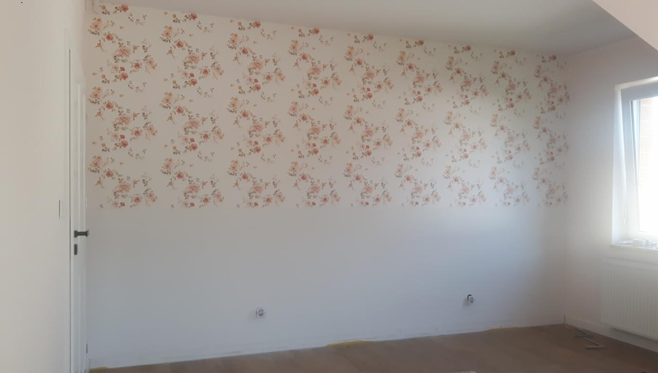 Ściana pokoju, na której górnej połowie widoczna jest tapeta w kwiaty, a dolna część jest biała, z widocznymi gniazdkami elektrycznymi i drzwiami po lewej stronie.