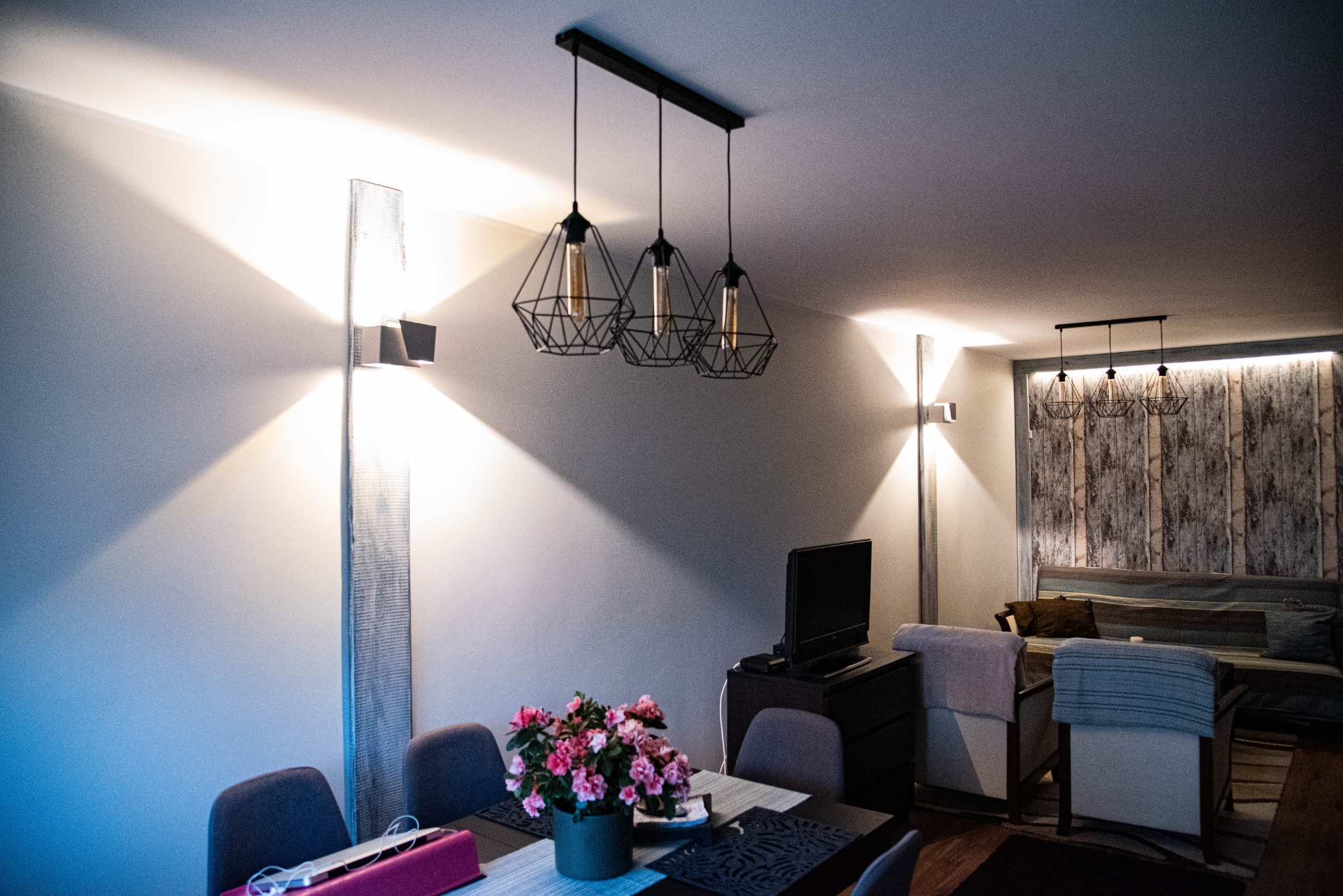 Wnętrze pokoju z nowoczesnym oświetleniem ściennym i wiszącymi lampami w stylu loft, kwiaty na stole, ściana pomalowana na jasny kolor.